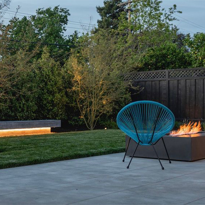 A modern hardscaping project with a large wooden bench lit underneath, tile terrace and fire pit.